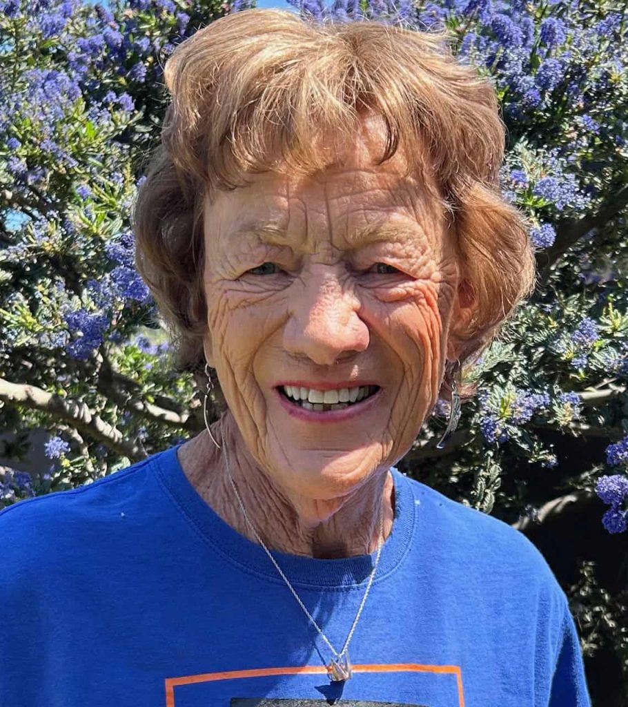 An older white woman with light brunette hair smiles at the camera. She is wearing a blue shirt, necklace and dangly earrings and is in front of a blue flowered blush outside.