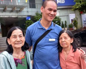 A Vietnamese man smiling and on either side of him are two elder Vietnamese women who were his former caretakers as a child.