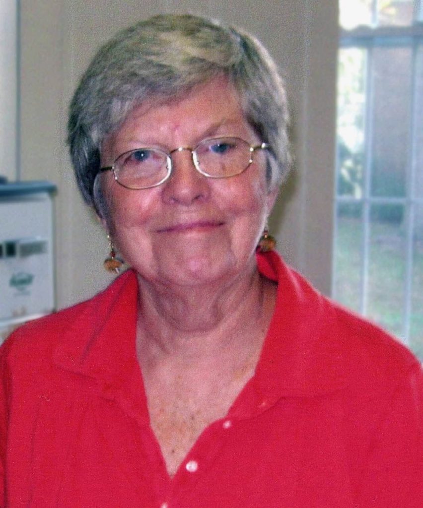 An older woman smiles softly at the camera. She has short grey hair, glasses, dangly earrings and is wearing a bright red polo shirt.