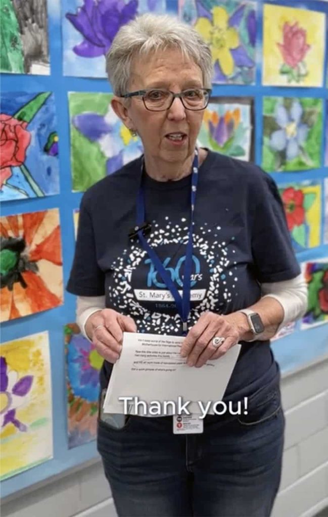 Phot of an older woman mid-sentence. She has short grey hair, glasses and is standing in front of a colorful art wall full of student drawings of flowers.