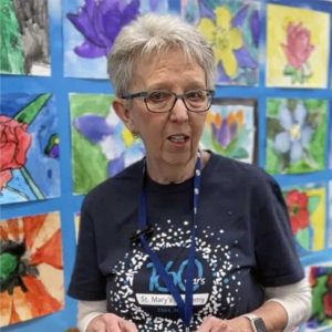 Phot of an older woman mid-sentence. She has short grey hair, glasses and is standing in front of a colorful art wall full of student drawings of flowers.