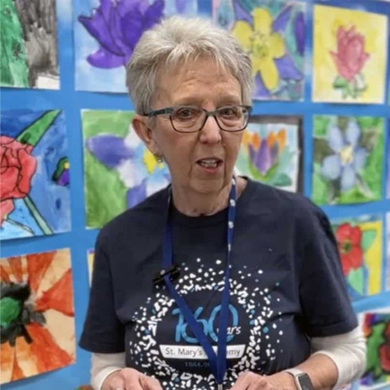 Phot of an older woman mid-sentence. She has short grey hair, glasses and is standing in front of a colorful art wall full of student drawings of flowers.