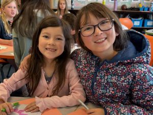 Two elementary students are shown smiling as they color prayer flags for peace.