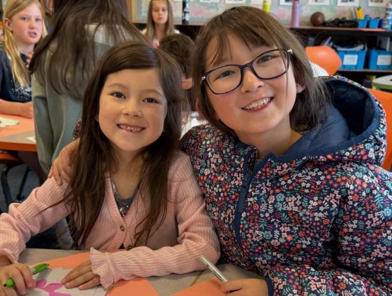 Two elementary students are shown smiling as they color prayer flags for peace.