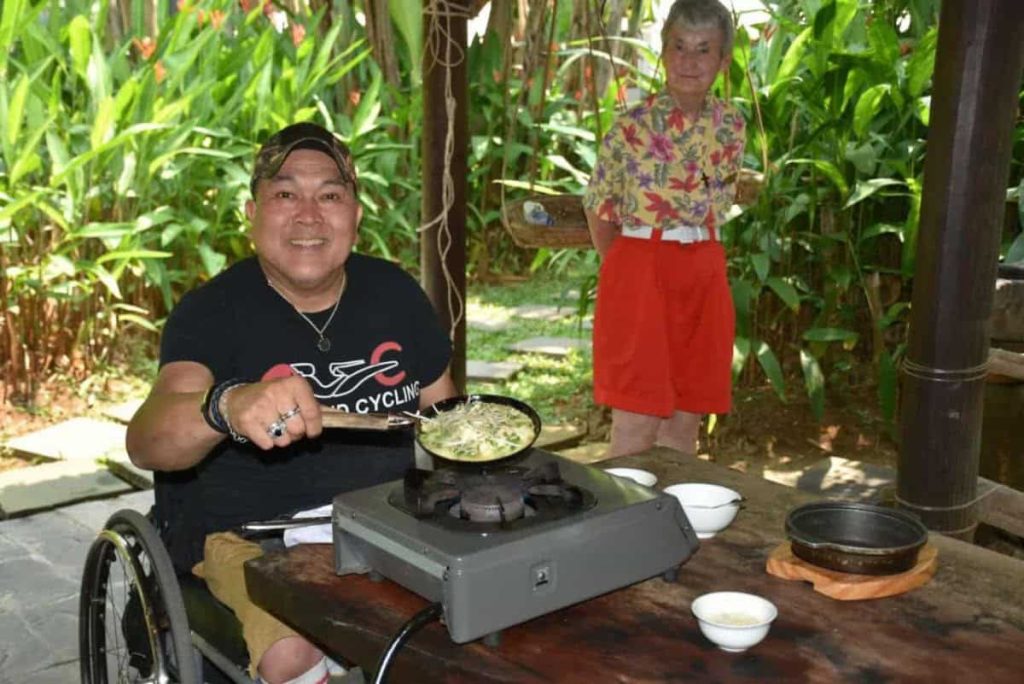 A man in a wheelchair smiles and shows the camera what he's cooking in an a skillet in an outdoor cooking school.