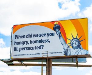 A billboard on a sunny day with the Statue of Liberty and bright orange and yellow rays behind her with the text: When did we see you hungry, homeless, ill, persecuted? Dominican Sisters of Peace and the Loretto Community