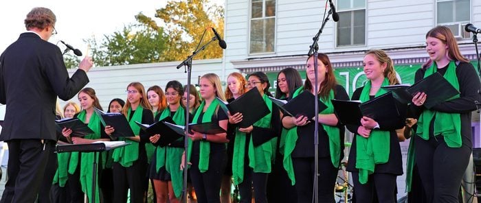 A man directing a choir of young women at an outdoor event. The choir singers are holding sheet music and wearing matching black outfits with green scarves.