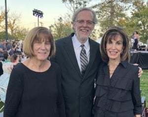 Two women and one man smile at an outdoor event in nice outfits. The man is wearing a suit and tie and both women are wearing black dresses.