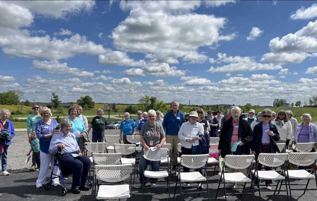 A group of people at an outdoor event standing around white chairs and smiling.