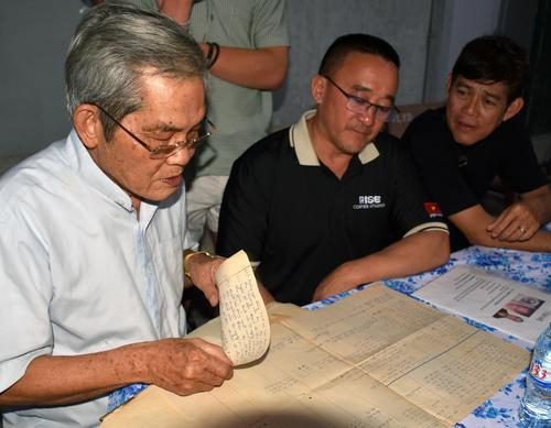 Three men sitting studying aged handwritten paper records at a table in a dimly lit room.