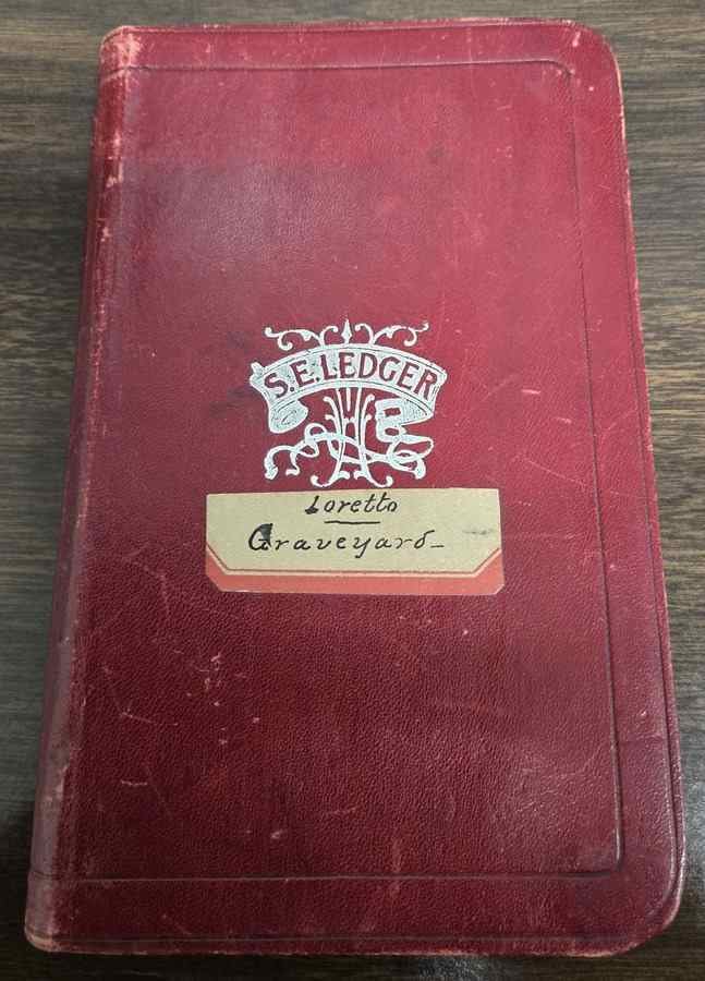 Worn red notebook with "S.E. Ledger" stamped on it and "Loretto Graveyard" handwritten on a label affixed to the front.