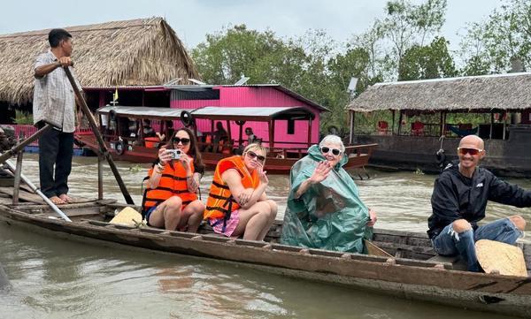 Four people sitting on a small boat and one man standing and rowing in a body of water in remote town with buildings with straw and grass roofs in the background. Two of the people sitting in the boat are wearing life jackets, and one is wearing a green plastic rain poncho.