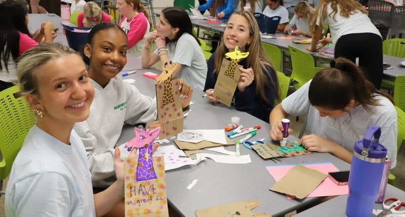 Five students sitting together working on craft projects at a school event full of other students working on crafts too.