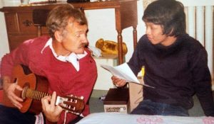 A man playing a guitar and a woman singing reading the lyrics on a paper. They are sitting on the floor together in a room with a wooden desk behind them.