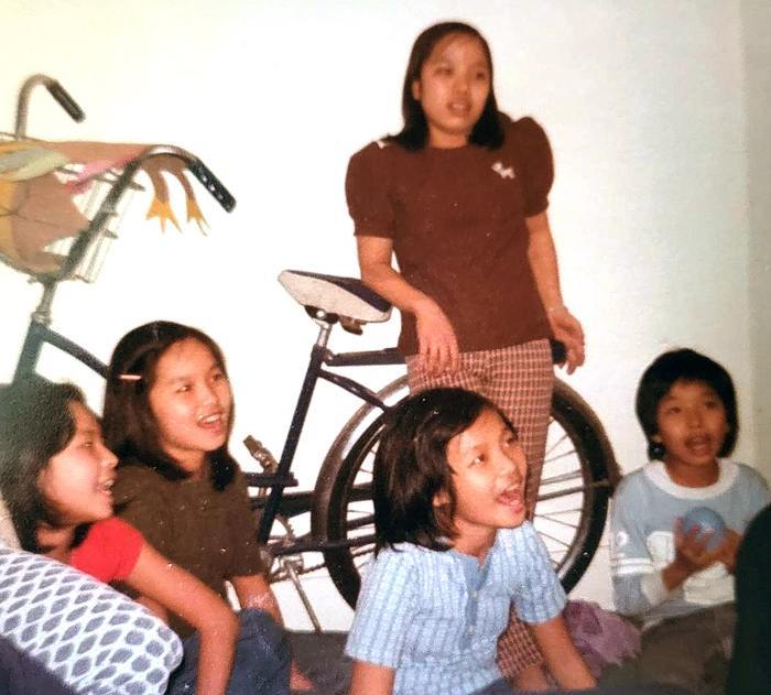 One young girl standing resting her elbow on the seat of a bicycle and four other children sitting and smiling at something out of frame.