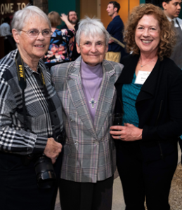 Three women standing together and smiling at at event wearing professional clothing.