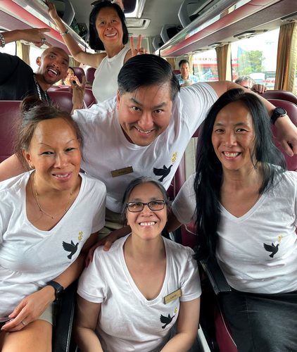 Six people smiling together, some wearing matching white t-shirts with matching bird graphics on a tourbus together.