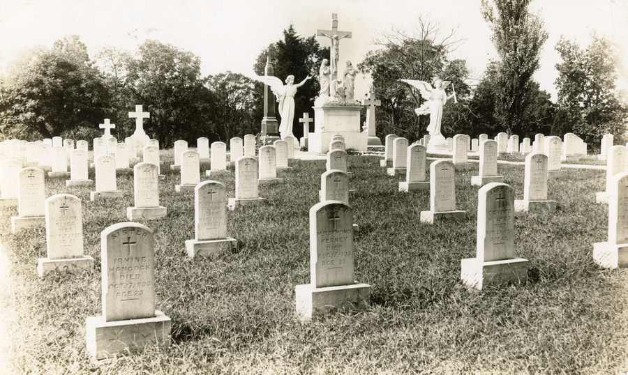 Archival photo of cemetery with rows of headstones, carved angels and a portayal of the crucifixion of Jesus.