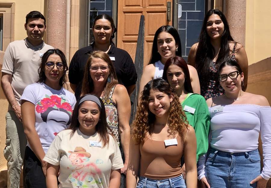Nine women and one man of different ethnicities pose in the sun for a group photo on the stairs leading up to a building.