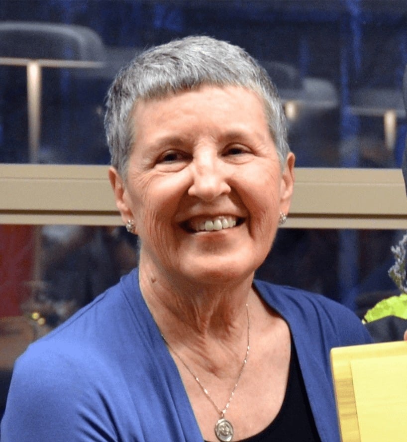An older white woman with short grey hair, silver earrings and necklace, with a blue cardigan smiles as she glances to the side.