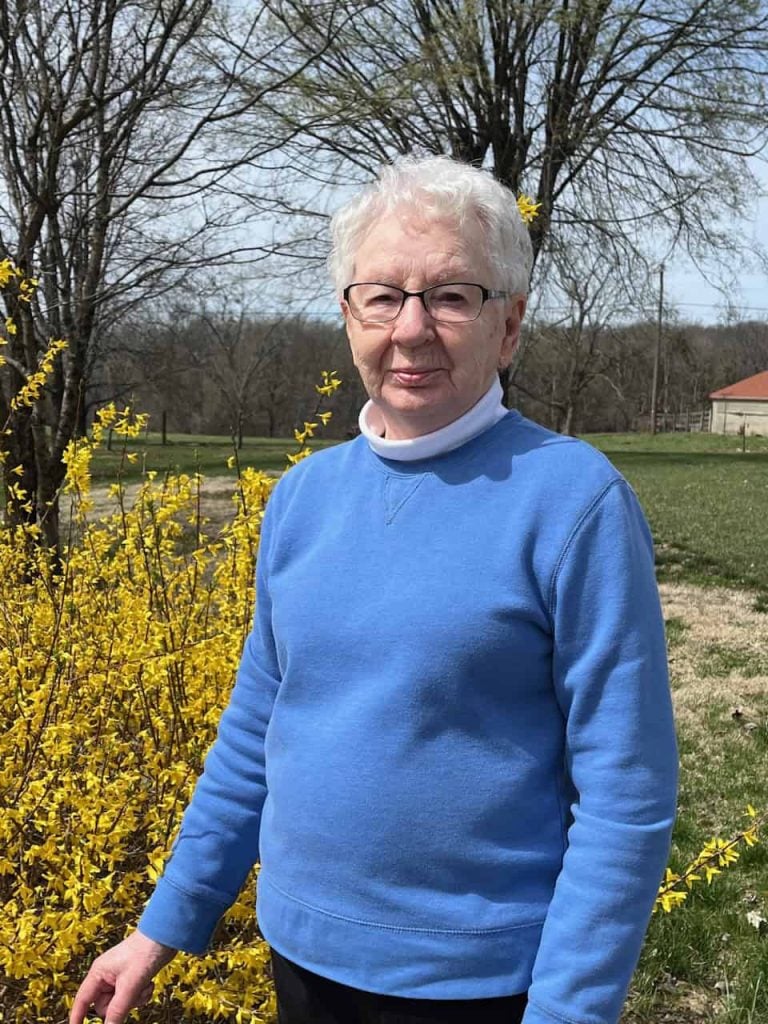 An older white woman with grey hair, glasses and a blue turtleneck with a white collar smiles softly outside in front of yellow flowers.