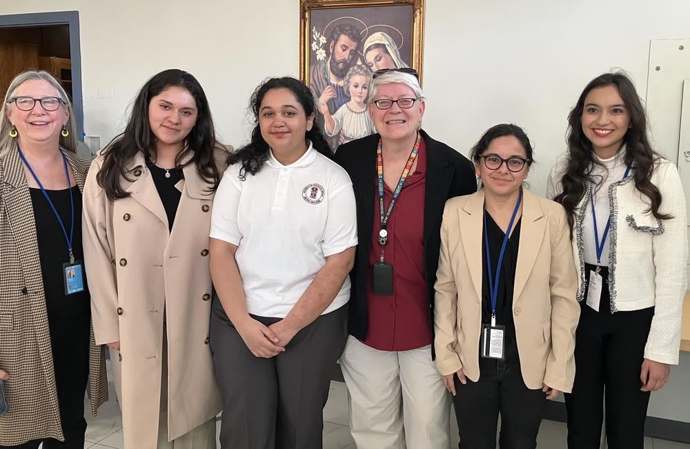 6 women smiling together for a picture indoors wearing business casual and matching lanyard passes.