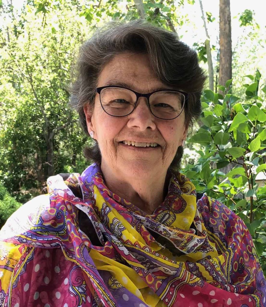An older white woman with brown and grey short wavy hair smiles. She is wearing large glasses and a colorful scarf covering her body in pink, purple, yellow and white colors.