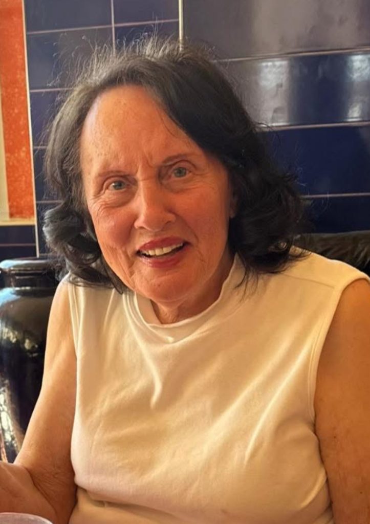 An older white woman smiling the camera, she has shoulder length dark brown hair and is wearing a yellow tank top.