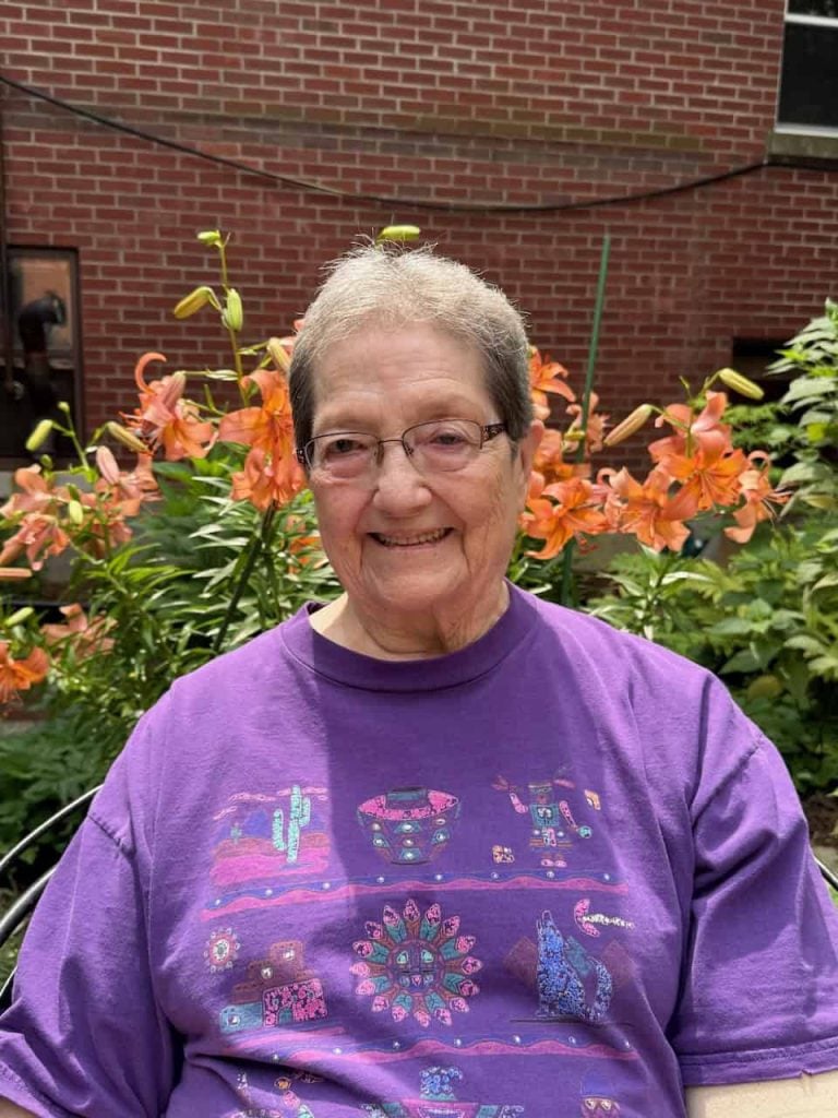 An older woman sitting outside smiling in front of blooming orange flowers. She has short grey hair, rectangular glasses and a purple shirt.