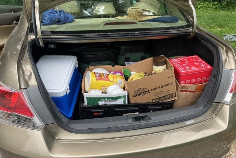 A look inside a trunk of a car prepped for a road trip. A large cooler and three boxes of food: lemonade, bananas, cookie boxes and more are visible in the trunk and a stack of woven sun hats rest in the back window.