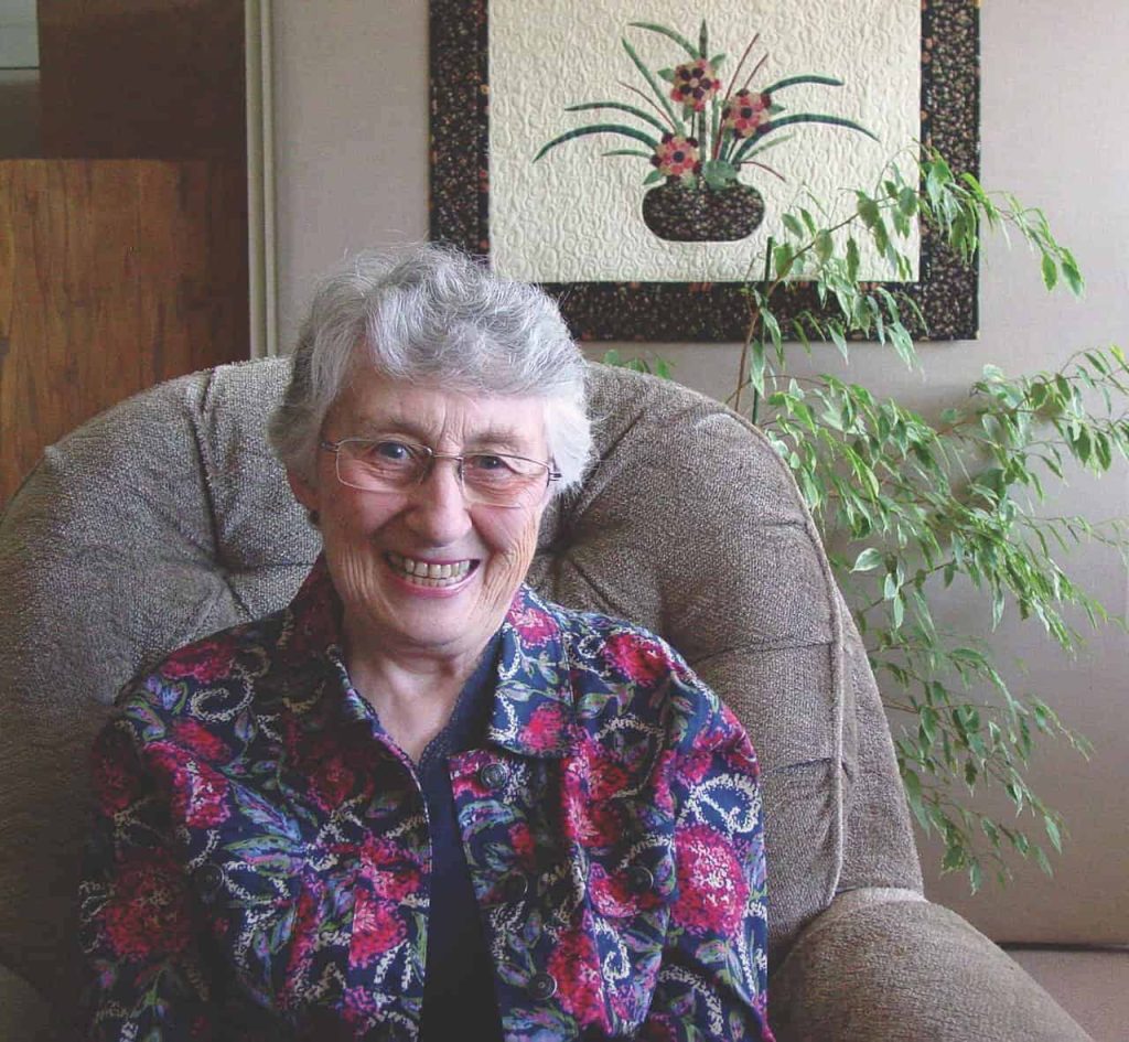 An older white woman smiling wide for the camera sitting on a grey chair. She has short wavy grey and white hair, rectangular glasses and a pink and purple floral jacket