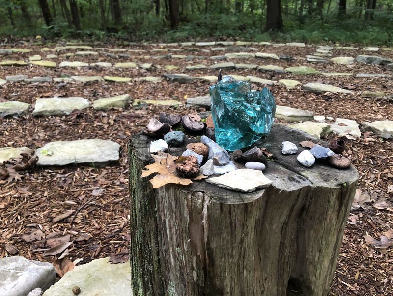 The center of a labyrinth of rocks on the forest floor with green trees behind it. In the center, there is a wooden stump with leaves and rocks placed on top.