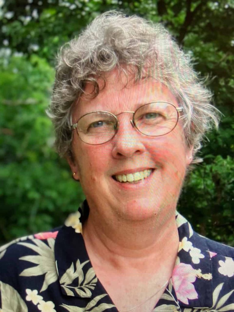 An older white woman with short grey curly hair smiling. She wears oval glasses, stud earrings and a floral shirt of green, pink and black.