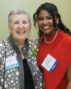 Two women wearing name tags and dress clothes smile together for a picture in front of a pale green wall.