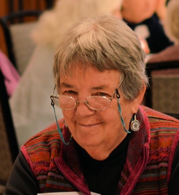 An older woman with shorter grey and white hair looks past the camera with a soft smile. She is wearing oval glasses with a blue glasses chain hanging down. She has dangle earrings and is wearing a striped red sweater.