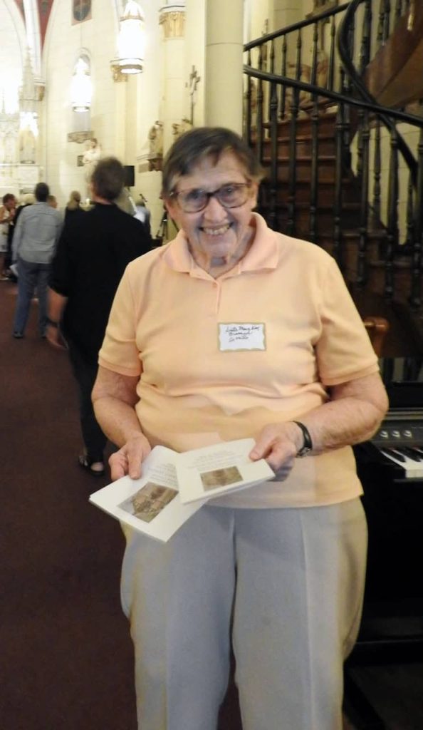 An older white woman with short brown hair wearing a light orange polo and grey pants in front of the Loretto staircase in New Mexico