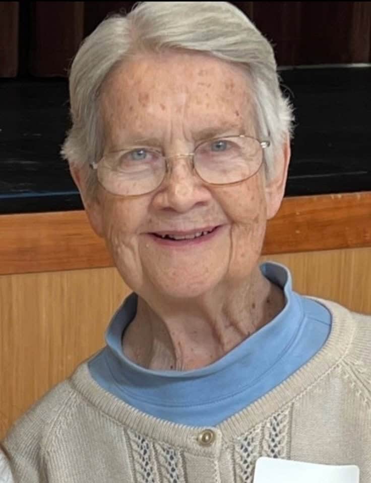 An older white woman with short grey and white hair smiles and looks at the camera. She has freckles on her face, is wearing oval glasses and a blue shirt with a white sweater over it