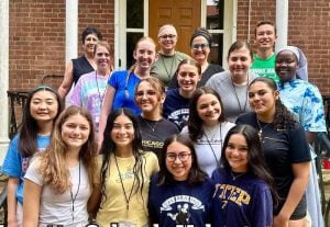 17 student and teachers gather together on stairs in front of a brick building and smile together for a group picture on a sunny day.