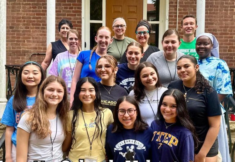 17 student and teachers gather together on stairs in front of a brick building and smile together for a group picture on a sunny day.