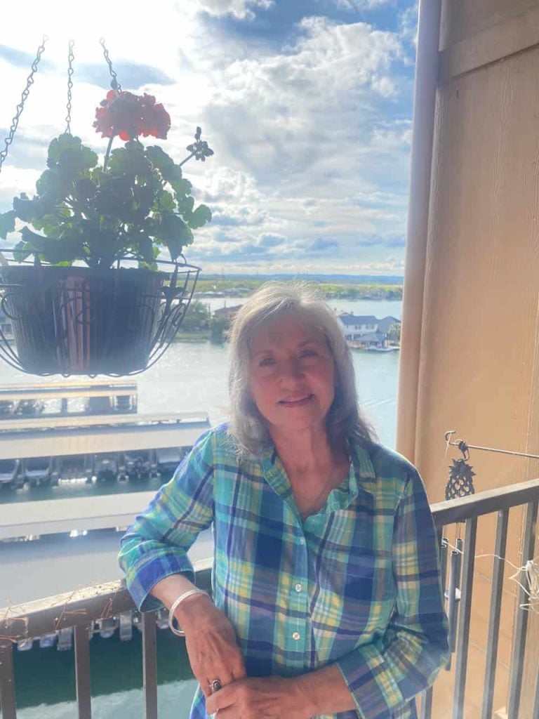 A middle aged white woman with medium length grey hair stands outside on a porch smiling wearing a blue, and yellow plaid shirt.