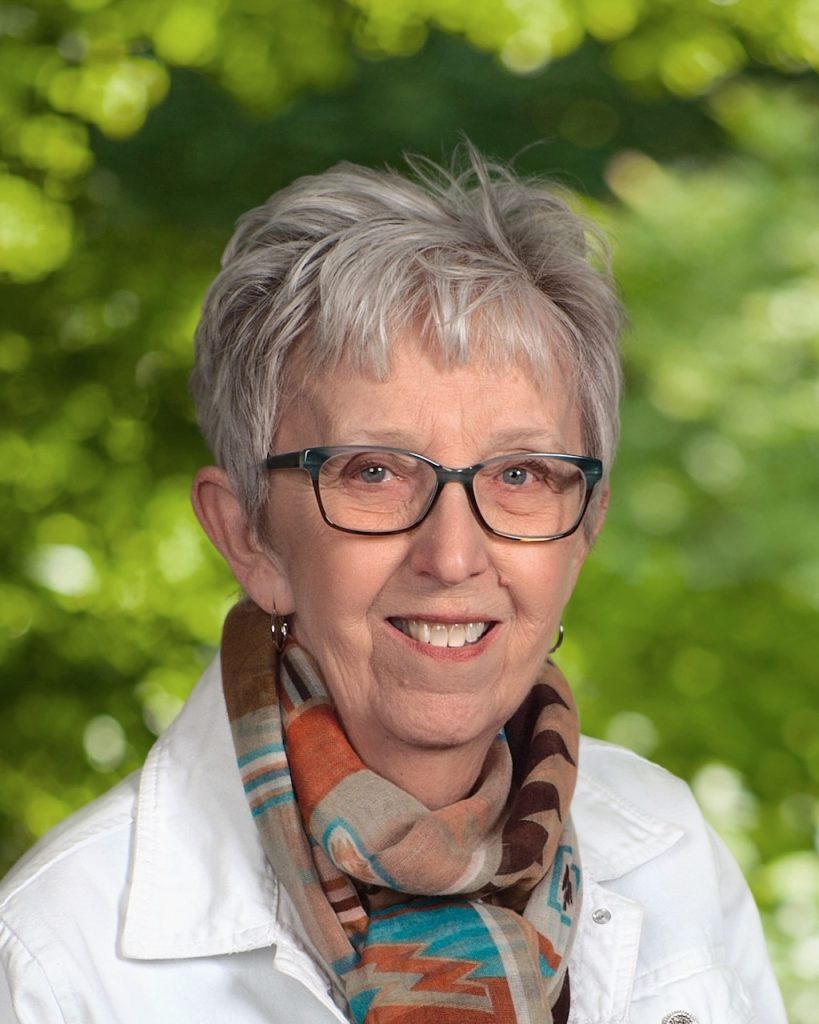 An older white woman with styled grey hair smiles for a photo. She has blue tinted glasses, small silver hoop earrings a white jacket and a colorful pattered scarf in blue, orange, brown and white.