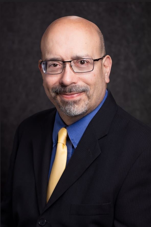 A middle aged bald white man with a rectangular glasses and a brown and white goatee smiles softly for a professional photo. He is wearing a black suit with a blue shirt and yellow tie underneath.