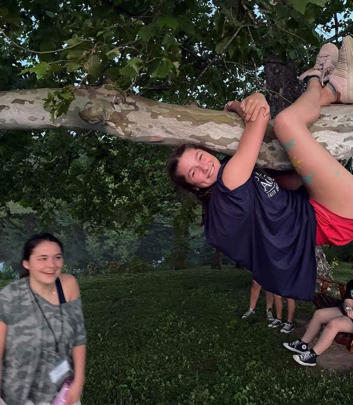 Two girls laughing outdoors at dusk while one hangs from a tree branch and the other stands beneath her.