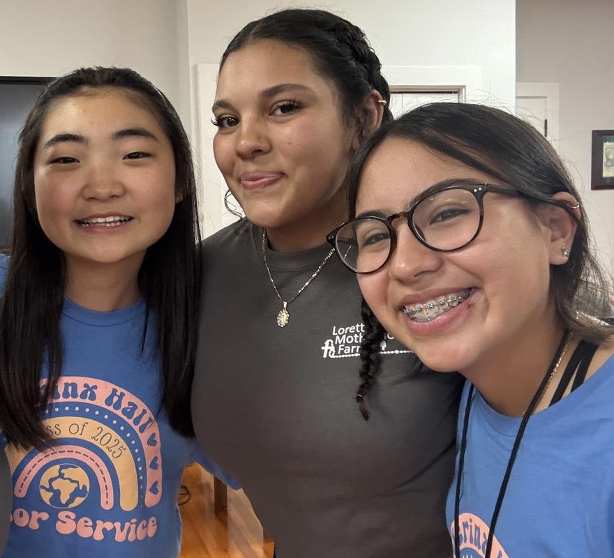 Three young girls smiling together for a group selfie indoors. Two of the girls are wearing matching blue and orange graphic T-shirts.