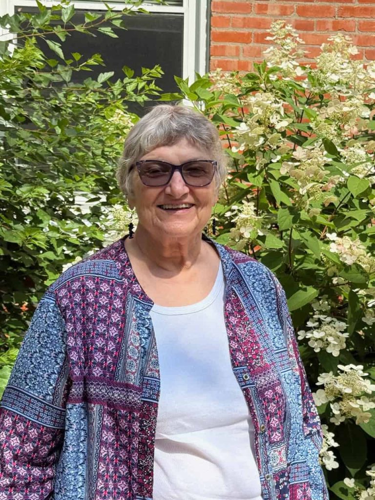 A portrait photo of an older white woman smiling in front of a flowering bush outside. She has short grey hair, is wearing translucent sunglasses, a white top and a patterned blue and purple cardigan.