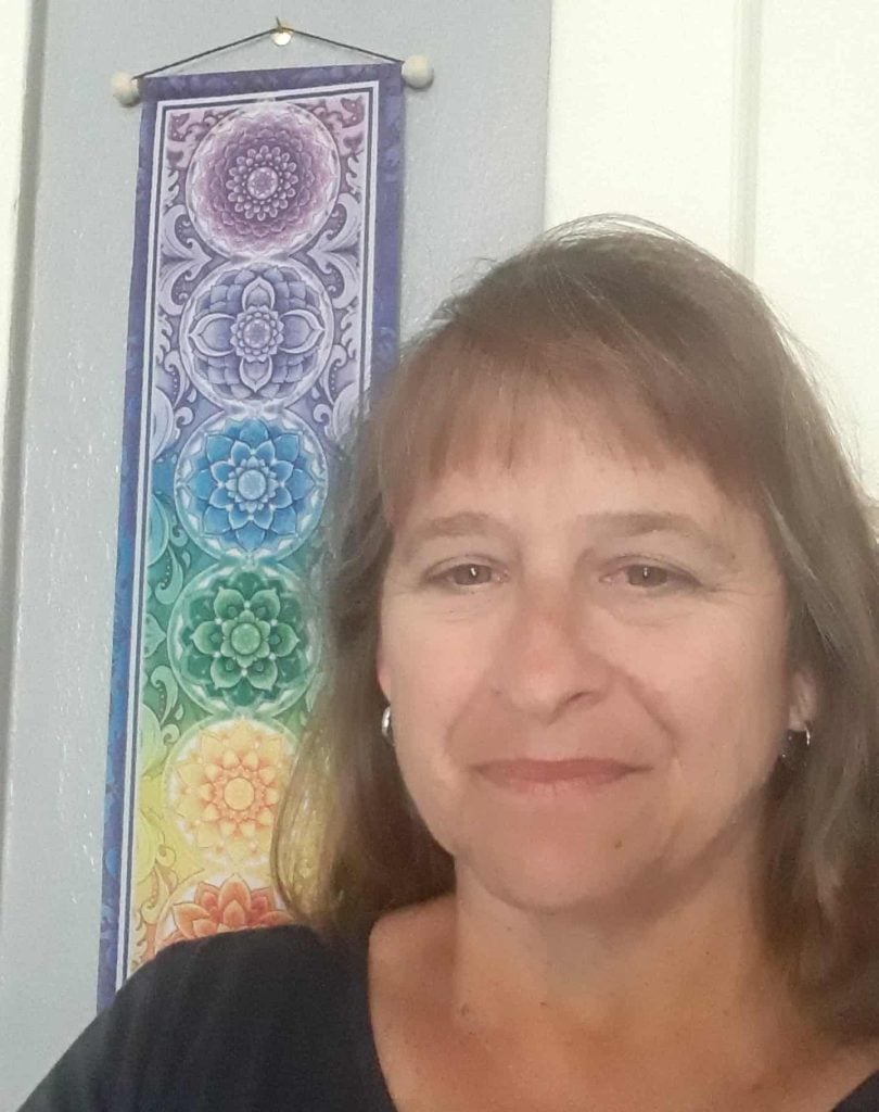 A middle aged woman with long brown hair and bangs smiles softly for a selfie. She is wearing small hoop earrings and a black shirt.