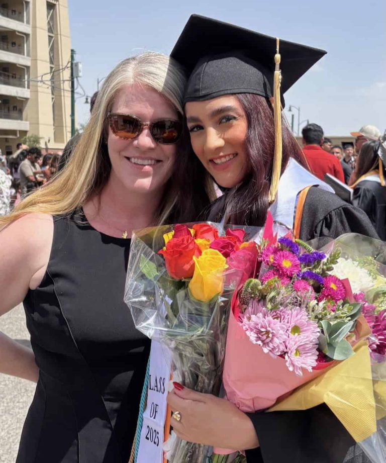 Two women, a White woman with long red hair and sunglasses and a Latina wearing a graduation cap and gown and stole reading "Class of 2025" and holding an armload of flowers, lean in together for a photo.