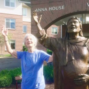 A white-haired woman raises her hand and looks to the sky with joy in an identical copy of the pose of the statue next to her.