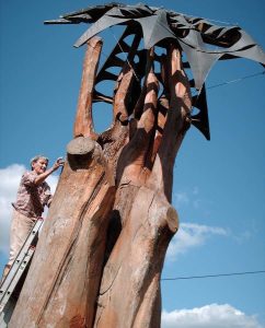 A woman stands on a ladder to work near the top of a huge sculpture. The base is shaped like a tremendous tree trunk, and a bird with black metal wings hovers over it.