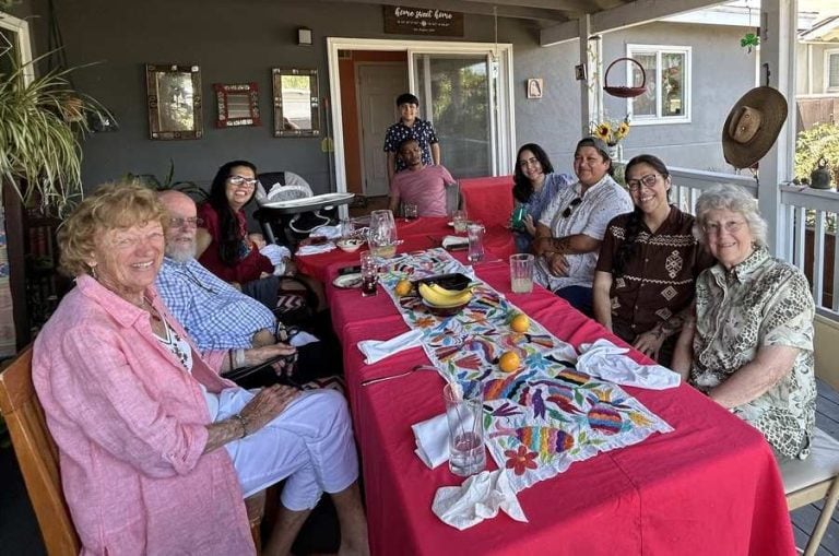 A group of people sit on a porch around a colorful table as part of a meeting discussion.